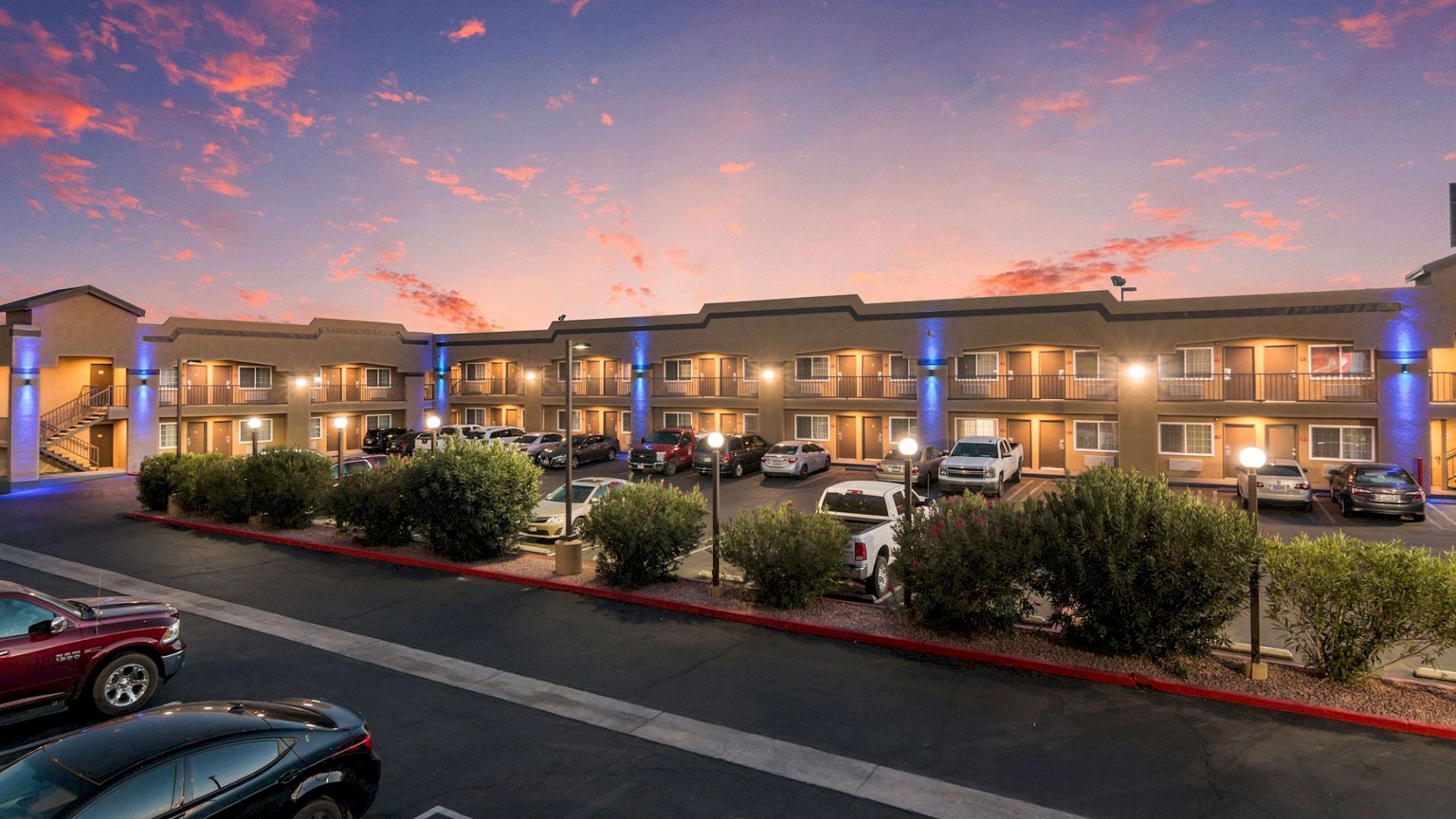 A two-story motel with parked cars and illuminated balconies under a vibrant sunset sky.
