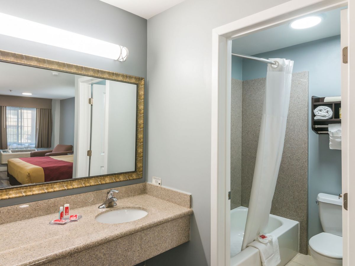 A hotel bathroom with a sink, mirror, and toiletries. Through an open door, a bathtub, toilet, and towel rack are visible. A bedroom is seen through another door.