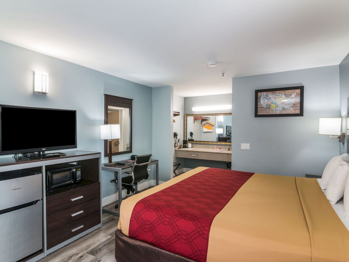 A modern hotel room with a bed, TV, mini-fridge, microwave, desk, wall art, and a mirror reflecting light from the bathroom area, ending the sentence.