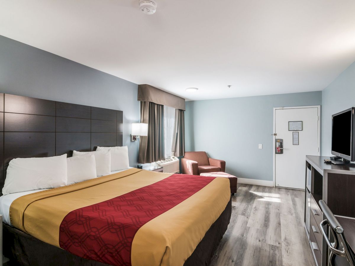 This image shows a hotel room with a bed, a TV, a chair, a desk, and a window. The room has a modern design and wood flooring.