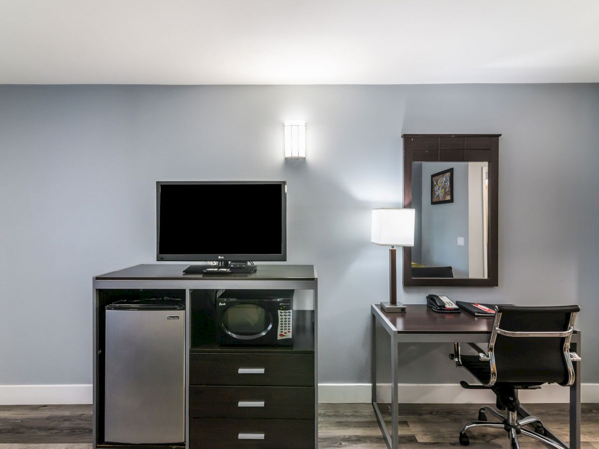 The image shows a hotel room setup with a TV atop a dresser containing a mini fridge and microwave, a desk with a chair, lamp, phone, and mirror.