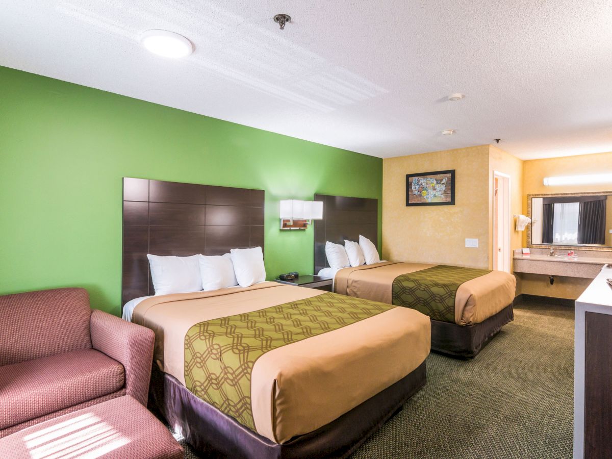 A hotel room with two double beds, a green wall, an armchair, a dresser, a vanity area with sink, and a framed picture on a yellow wall.