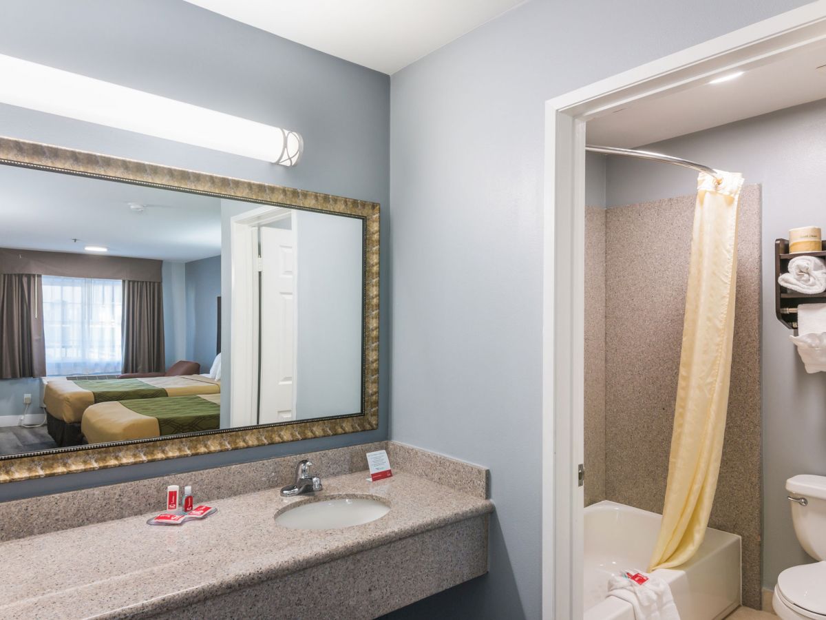 This image shows a hotel bathroom with a sink and large mirror, an adjacent shower with a yellow curtain, and a view of the bedroom in the reflection.