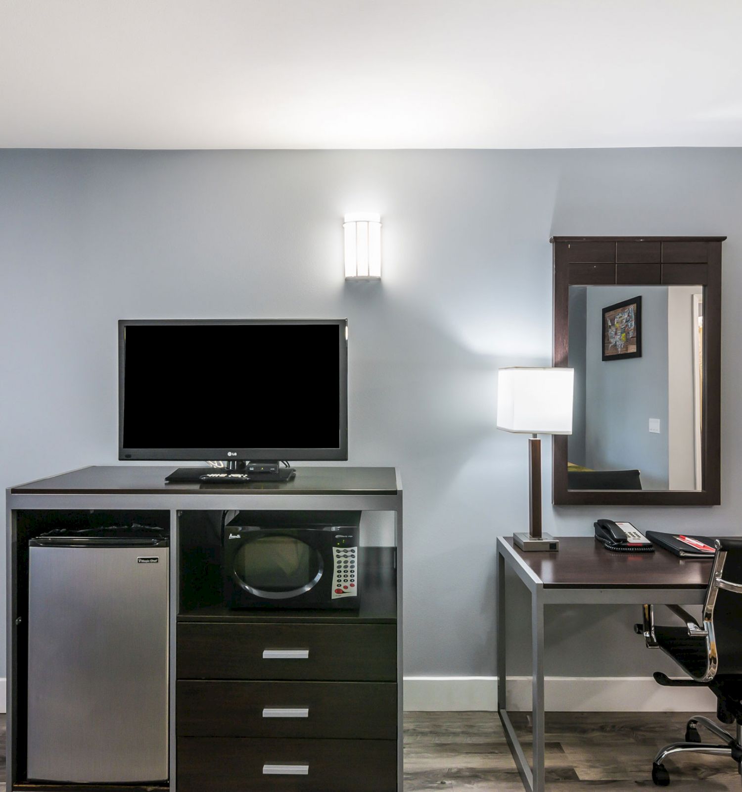 A hotel room corner with a TV, fridge, microwave on a cabinet, a desk with a chair, lamp, phone, and a mirror on the wall.