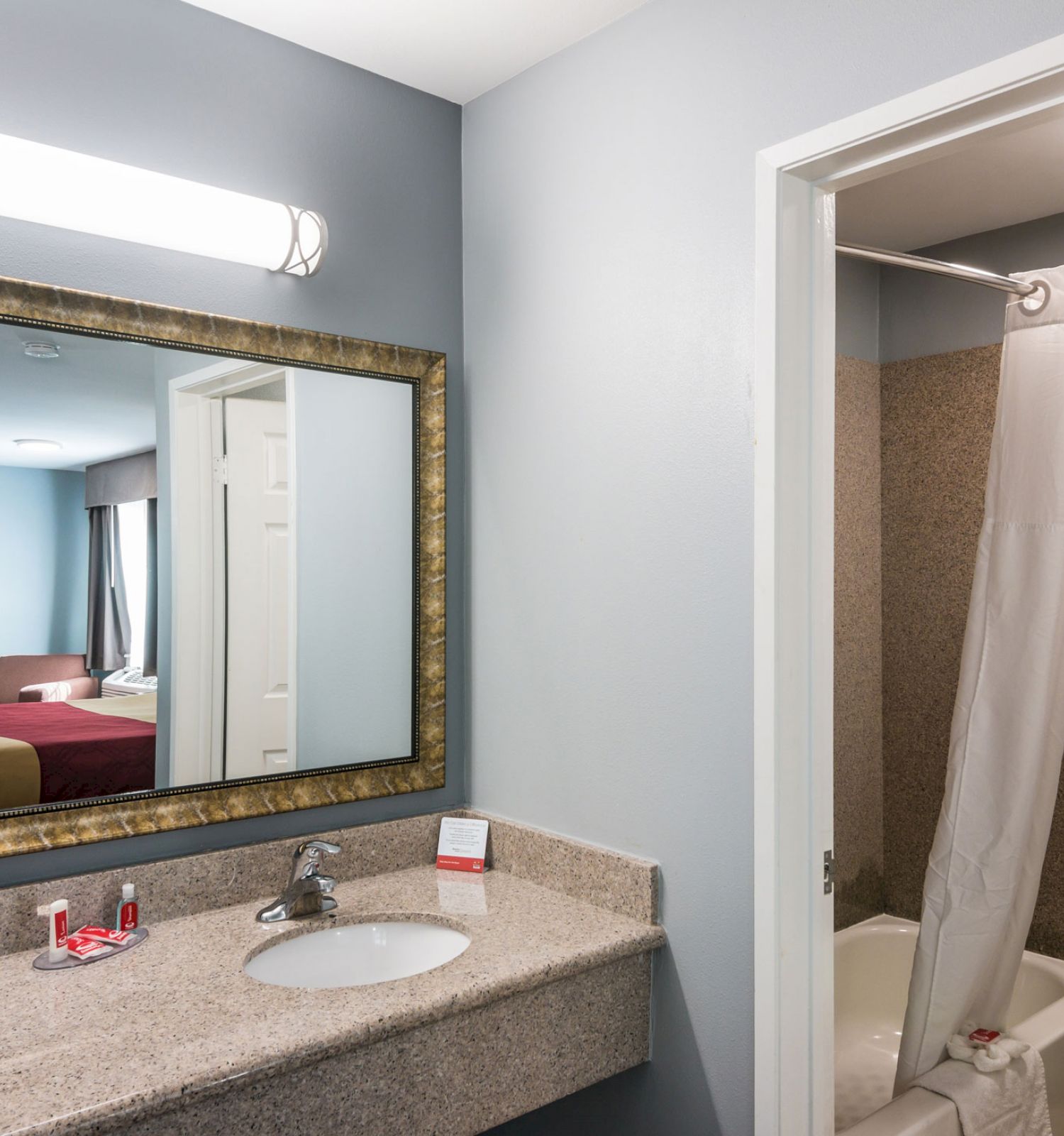 The image shows a hotel bathroom with a large mirror over a sink and a view into the bedroom. The shower has a curtain partially open.