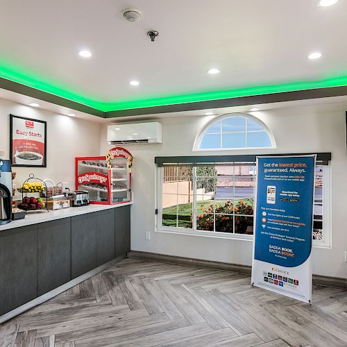 A small lobby area with beverage machines, snacks, a TV, and informational signage, under a ceiling with green lighting.