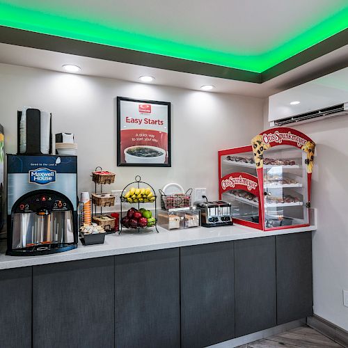 A small breakfast bar features juice and coffee dispensers, a fruit basket, a toaster, and a popcorn machine under green-lit ceiling lights.