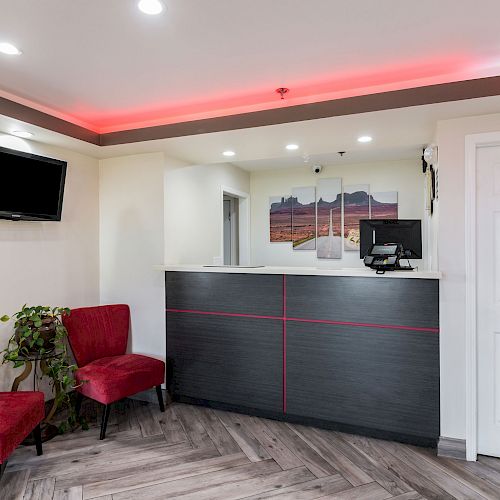 The image shows a modern lobby with red-accented ceiling lights, a reception desk, two red chairs, a wall-mounted TV, and some decorative plants.