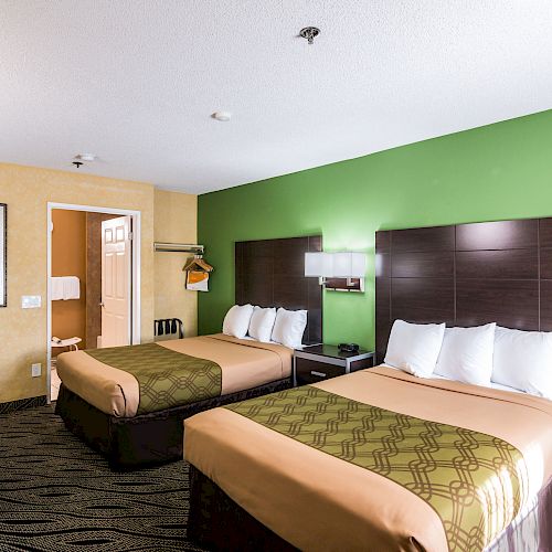 A hotel room with two double beds, a green accent wall, a wall-mounted TV, and a closet with hangers and an iron. The bathroom door is open.