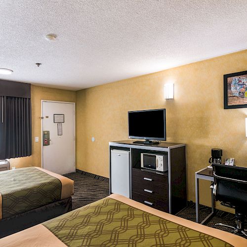 A hotel room with two beds, a TV, a desk with a chair, a fridge, and a window. The wall has a painting, and there's a lamp on the desk.