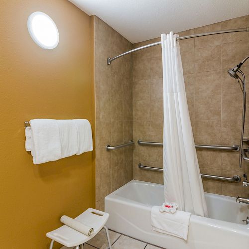 The image shows a bathroom with tan walls and a beige tiled bathtub area, featuring a shower curtain, towel rack, and a small white stool.