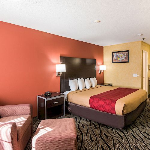 This image shows a hotel room with a bed, an armchair, a nightstand, lamps, and a vanity area with a sink and mirror.