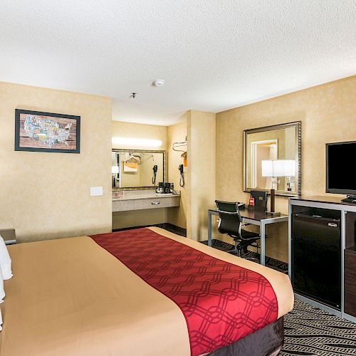 This image shows a hotel room with a bed, TV, microwave, desk, chair, and a vanity area with a mirror and sink.