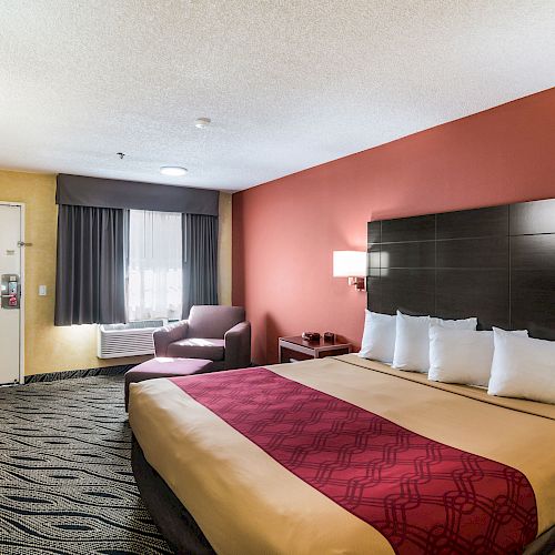 A hotel room with a king-size bed, red and yellow bedding, a TV, an armchair, a window with curtains, and a door. Modern décor and carpet flooring.