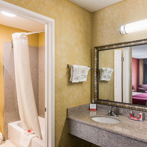 The image shows a hotel bathroom with a sink, large mirror, towels, and a shower-tub combo. A glimpse of the bedroom is visible through the mirror.