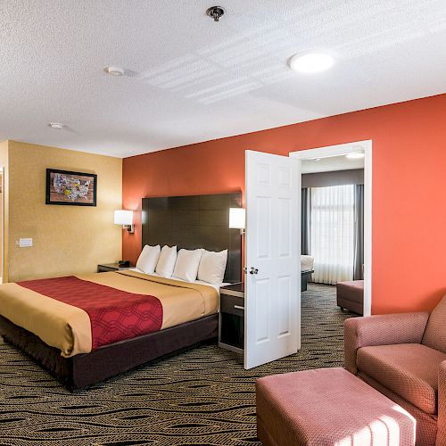 A hotel room with a king bed, an open door to an adjacent room, a bathroom sink, wall art, and a seating area with a chair and ottoman.