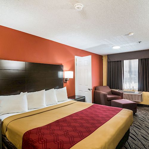 A hotel room with a king-size bed, red accent walls, nightstand, armchair by the window, and a closed door leading to the adjacent room or bathroom.