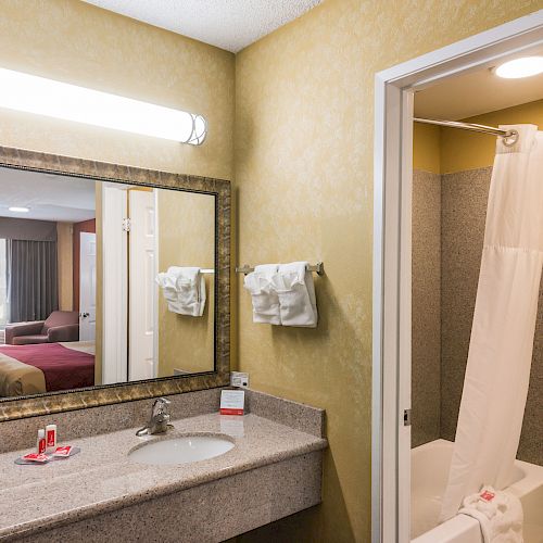 A hotel bathroom with a sink and large mirror, next to a shower and toilet, with a view of the bedroom in the reflection.