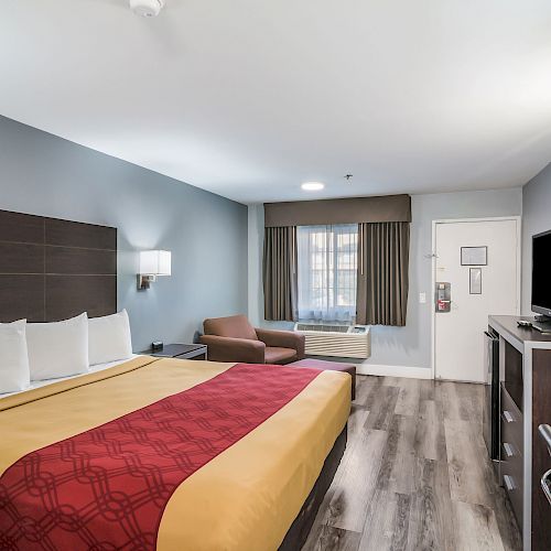 A hotel room with a large bed, TV, desk, chair, and a window. The bed has white pillows, and the room has modern furnishings.