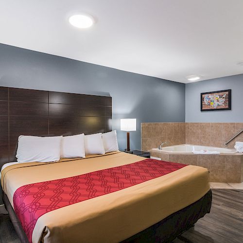 The image shows a modern hotel room with a king-size bed, a jacuzzi tub, bedside tables, lamps, and a sink area in the background.