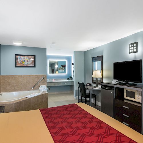 A hotel room with a bed, TV, microwave, refrigerator, a lamp, a desk with chair, and a bathtub with a grab bar.