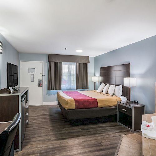 A hotel room with a bed, desk, TV, lamp, and an open area with a bathtub, featuring modern furniture and a neutral color scheme.