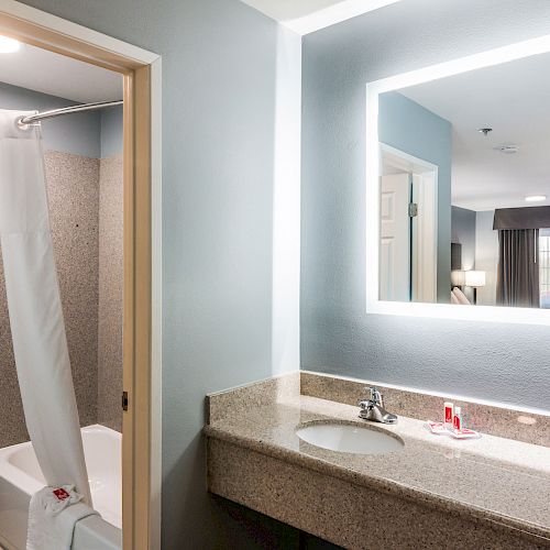 A well-lit hotel bathroom with a large mirror, sink, and granite countertop. The shower curtain is partially drawn open, revealing a bathtub and toilet.