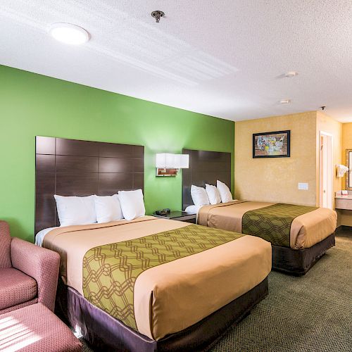 A hotel room with two double beds, a green accent wall, a cushioned chair, a vanity area with a sink, and framed artwork on the wall.