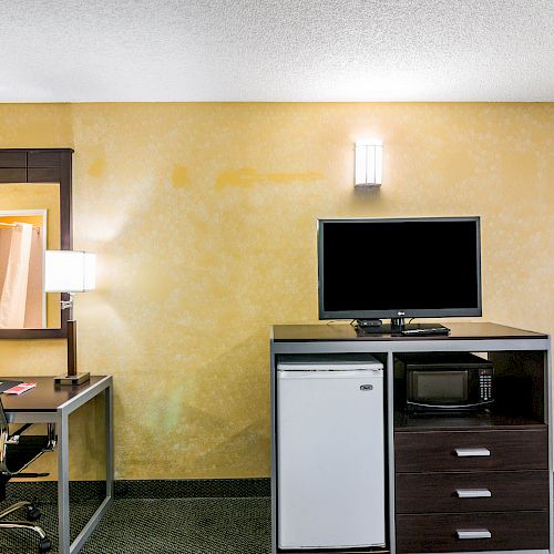 A hotel room with a desk, chair, lamp, mirror, TV, mini-fridge, microwave, drawers, and yellow walls.