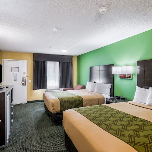 A hotel room with two double beds, a TV on a stand, a chair, a green accent wall, and a window with dark curtains.