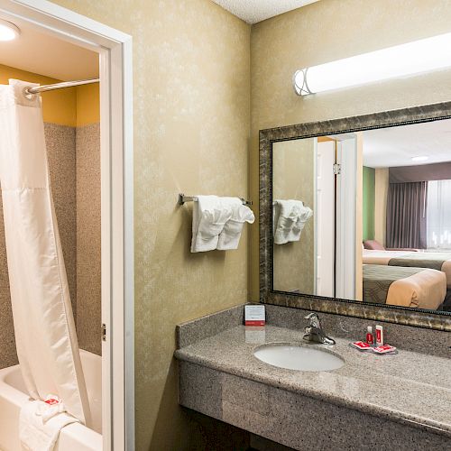 The image shows a hotel bathroom with a shower and toilet, a large mirror above the sink, and a glimpse of the room with a bed outside.