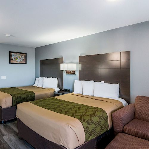 This image shows a hotel room with two double beds, a brown armchair, a vanity area with a sink, and a piece of framed artwork on the wall.