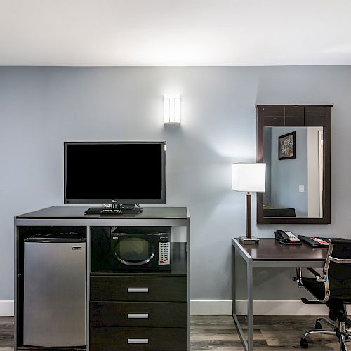 The image shows a hotel room setup with a TV, mini-fridge, microwave, desk, chair, desk lamp, and wall-mounted mirror.