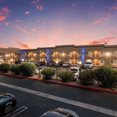 The image shows a two-story hotel or motel with several cars parked in front, surrounded by bushes, all set against a colorful sky at sunset.