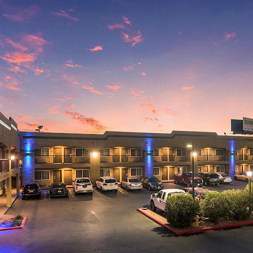 The image shows a two-story motel at dusk with cars parked in the lot, lit by building lights, and a vibrant sunset sky in the background.