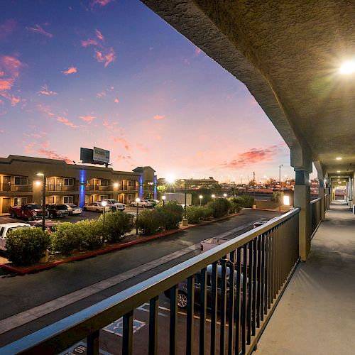 A motel exterior at sunset, featuring parked cars, illuminated buildings, and a walkway on the upper floor.