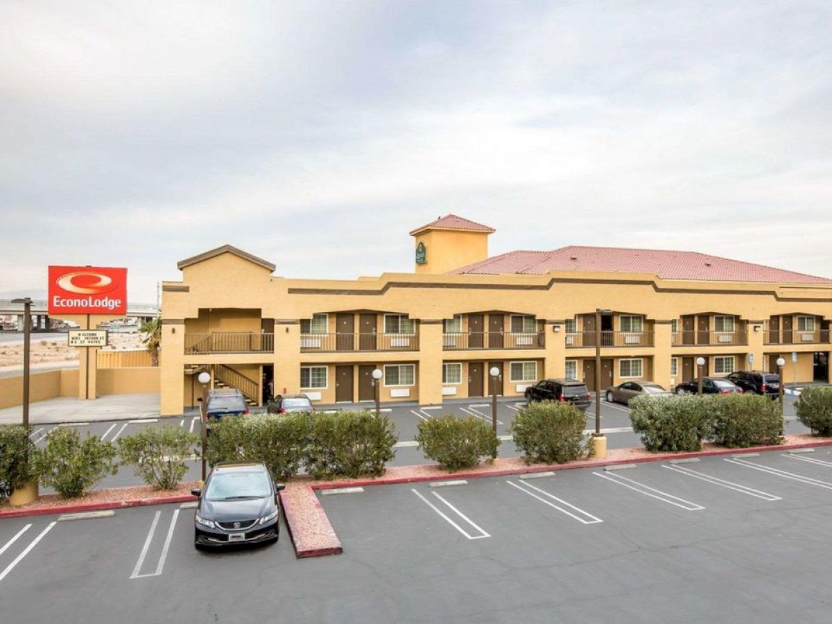 A two-story Econolodge motel with a red roof, several cars parked, and greenery in the parking lot under a cloudy sky ends the sentence.