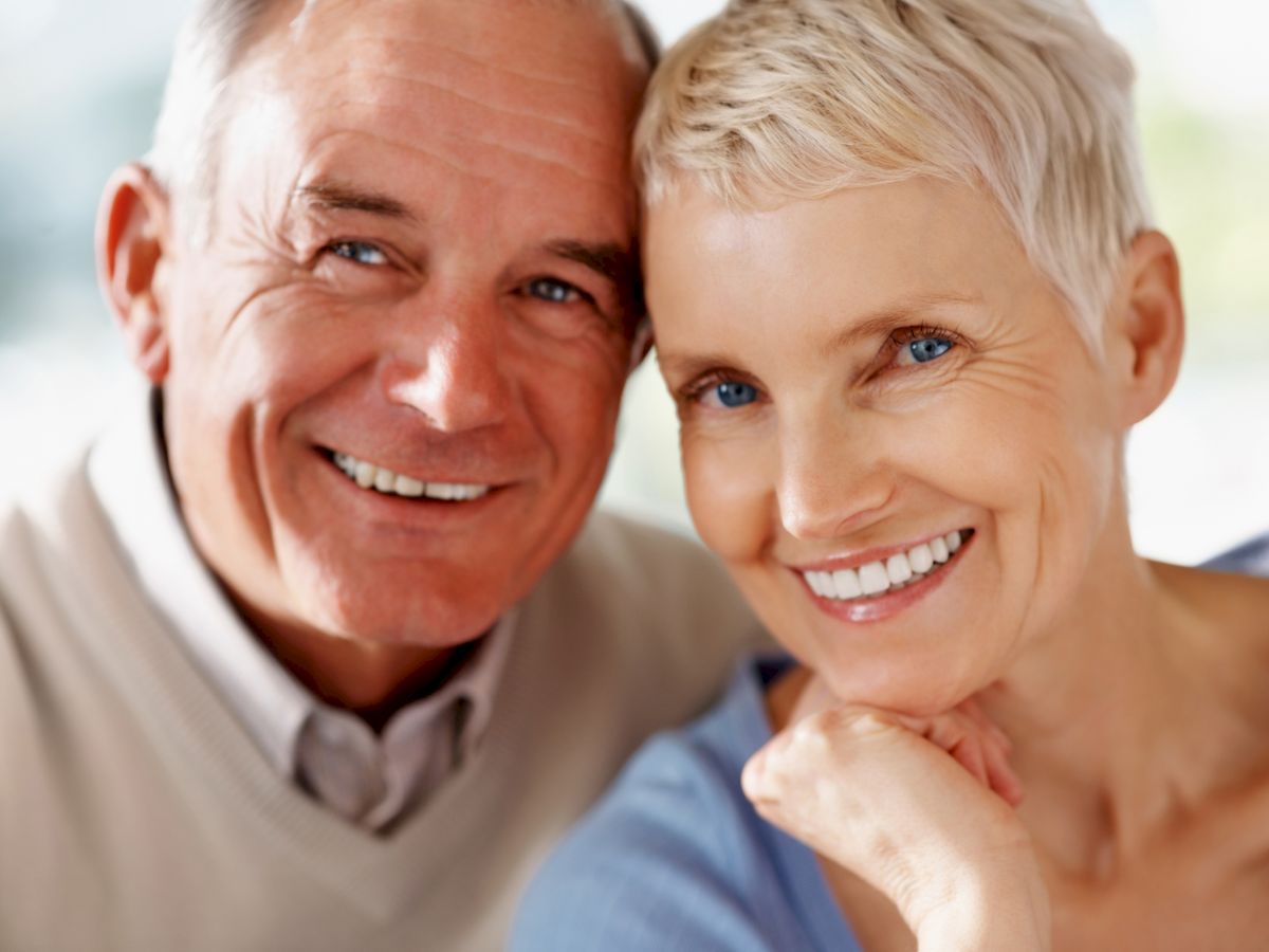 A smiling elderly couple is pictured, with the man wearing a beige sweater and the woman in a light blue top, both looking directly at the camera.