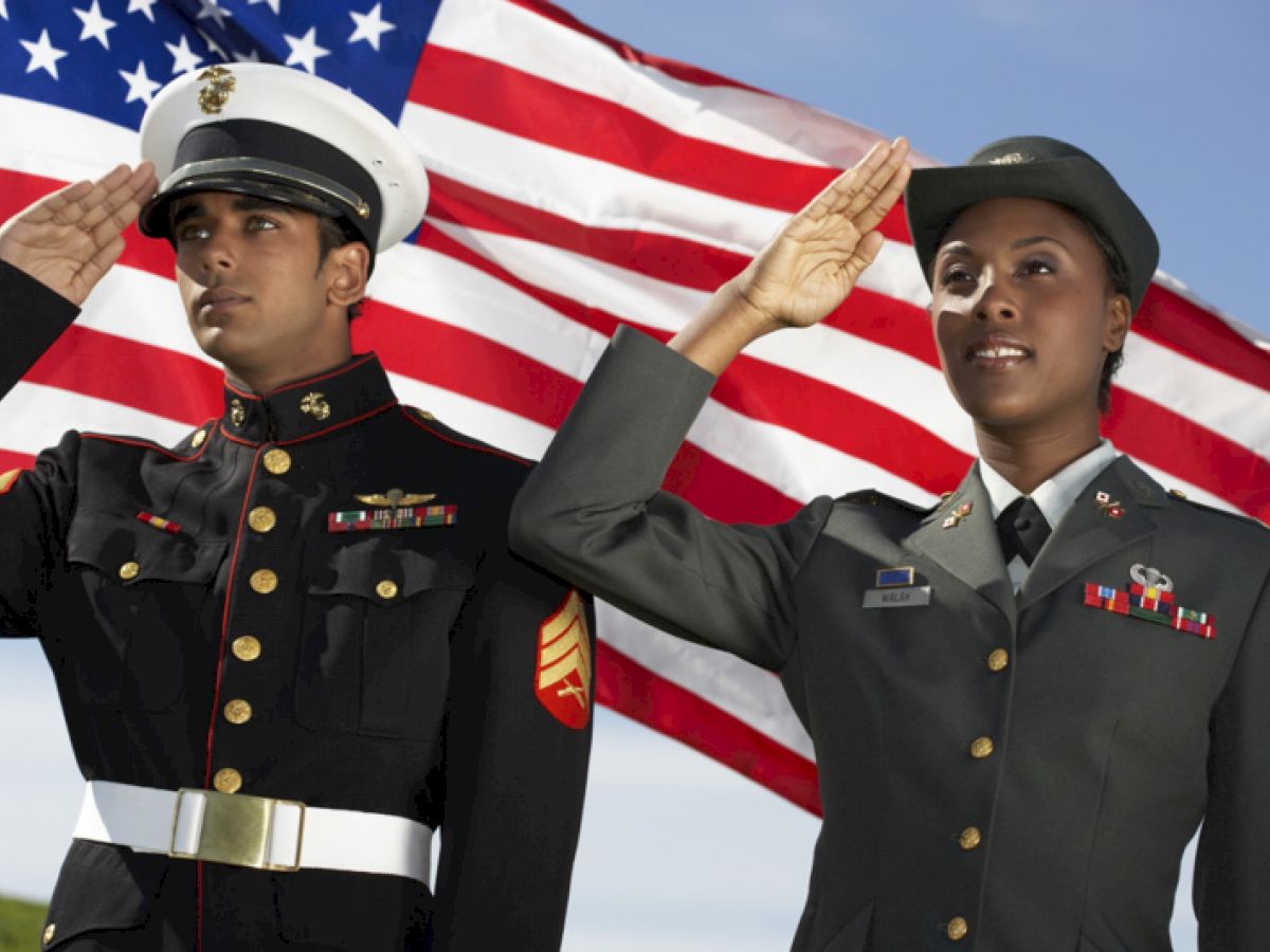 Two individuals in military uniforms salute in front of an American flag, representing dedication and respect.