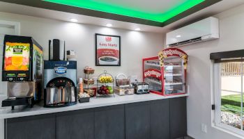 A small breakfast area featuring a juice dispenser, coffee machine, fresh fruits, a toaster, and a heated display case under green accented lighting.