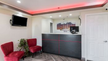 A modern reception area with a counter, computer, red chairs, wall-mounted TV, a plant, and a three-piece wall art, under recessed lighting.