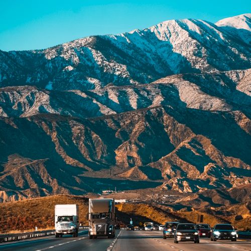 A highway with vehicles, including trucks and cars, set against a backdrop of large mountains with patches of snow under a clear blue sky.