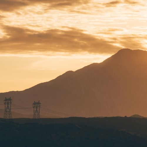 A mountain landscape during sunset with power transmission towers in the foreground and a sky filled with clouds, creating a serene scene.