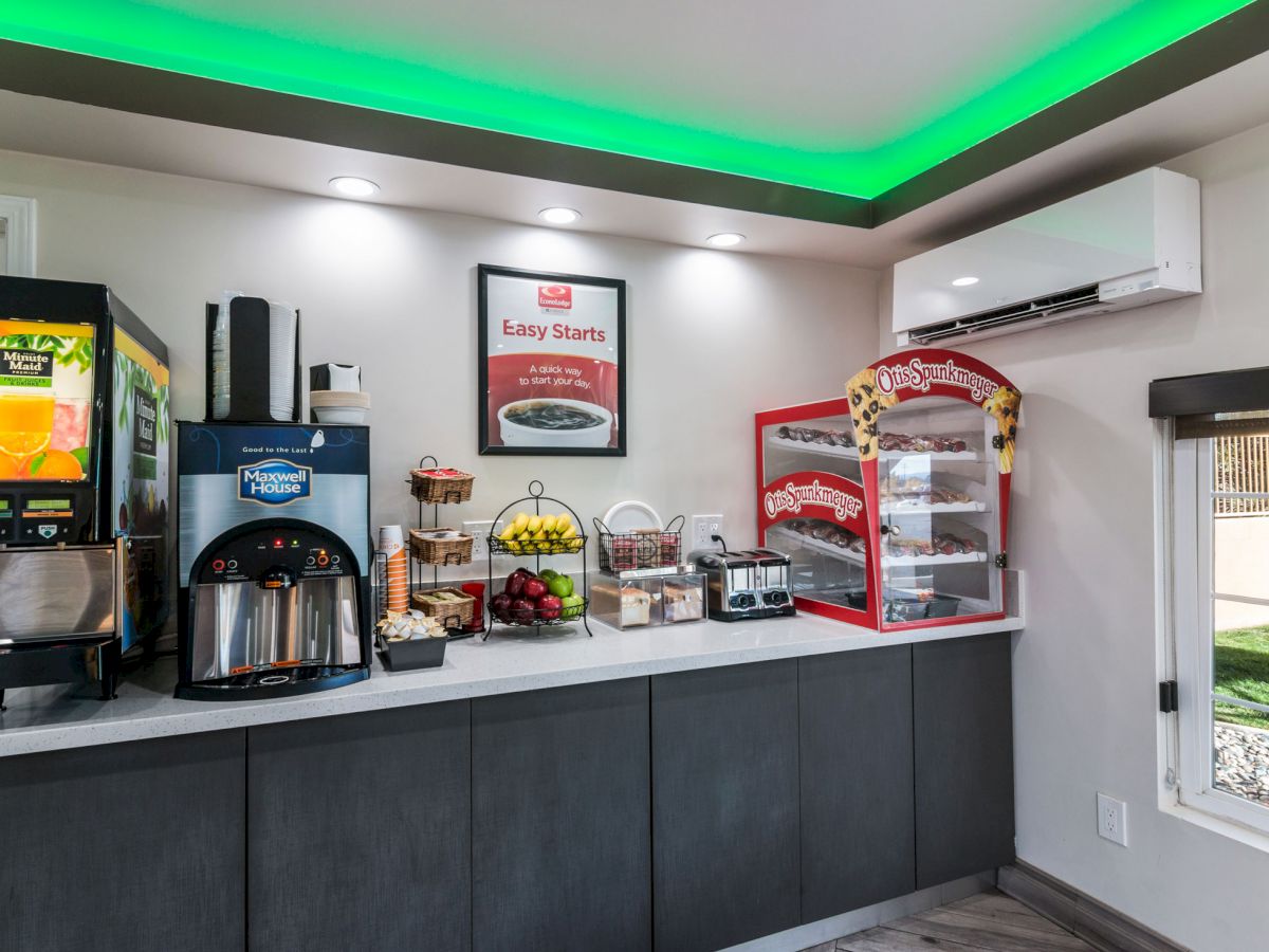 The image shows a small self-serve breakfast area with a juice dispenser, coffee machine, fruit, pastries, a toaster, and a bright green ceiling light.