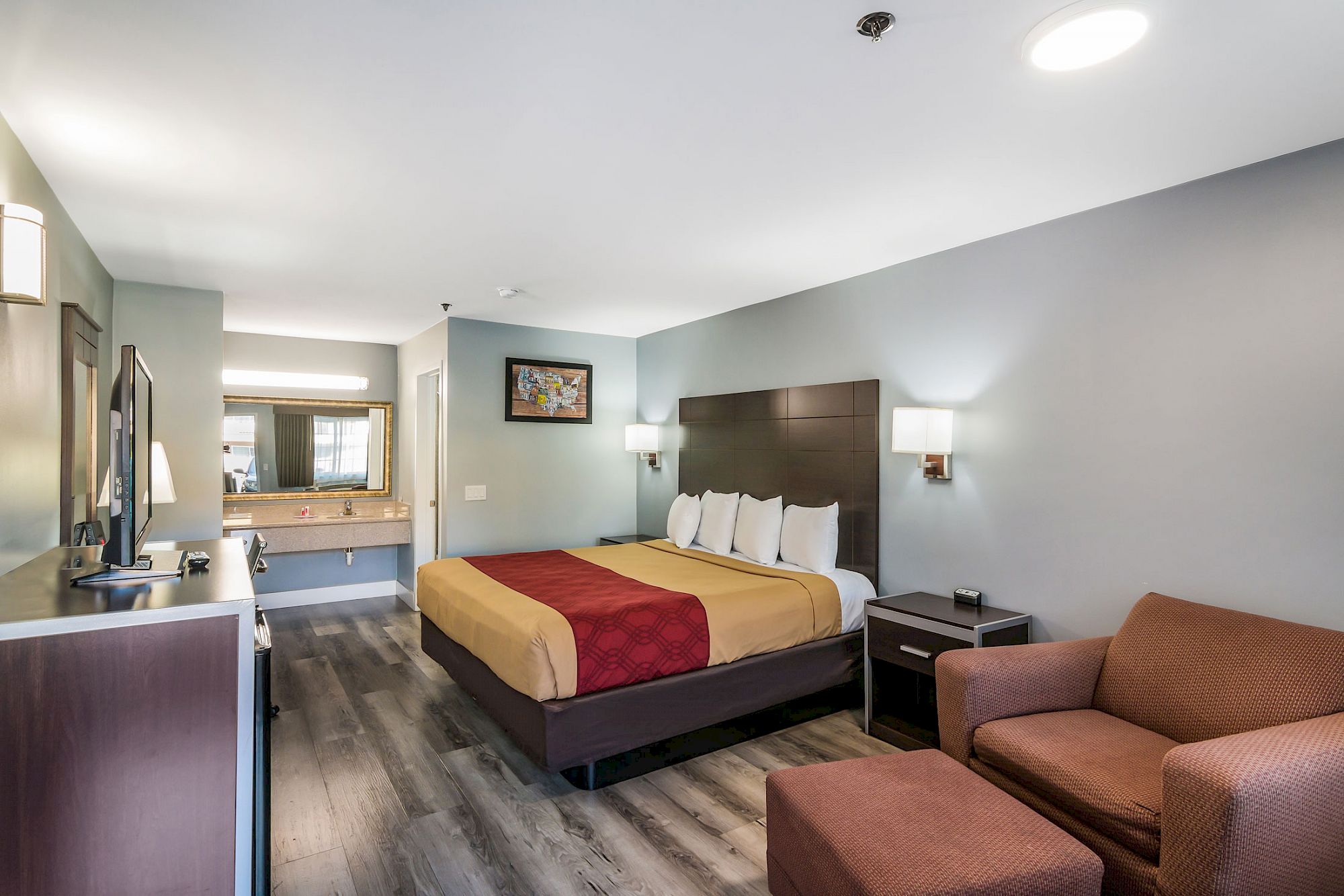 A hotel room featuring a double bed with yellow and red bedding, a chair with ottoman, a TV, a sink, and a small bathroom.