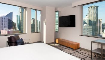A modern hotel room with a bed, chair, wall-mounted TV, and large windows showcasing a cityscape with tall buildings ending the sentence.