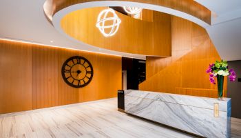 A modern lobby with a stylish reception desk, large clock on the wall, wooden paneling, and a curved staircase with contemporary lighting fixtures.