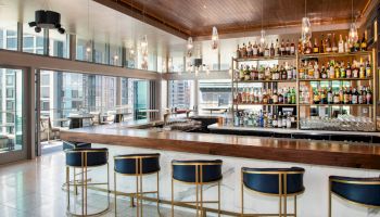 A modern bar with a wooden counter, stylish bar stools, and an extensive selection of liquor bottles is set against a backdrop of large windows.