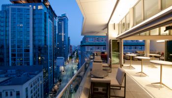 A modern balcony overlooking a cityscape at dusk, with glass buildings, street view, tables, and chairs, creating a stylish urban environment.