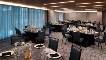 The image shows an elegant dining area set for an event with round tables, black chairs, glassware, and table settings under modern ceiling lights.